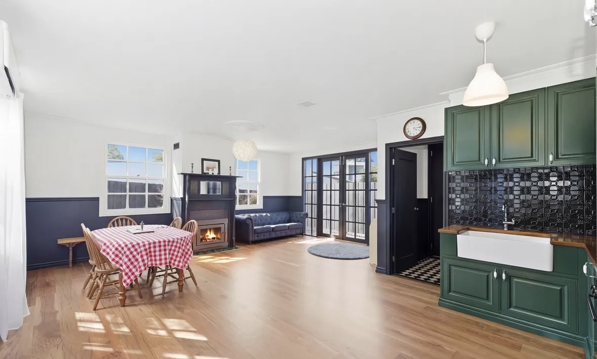 The finished living and dining room, with timber floors, an open fire and a dark green kitchen with a pressed-metal splashback.